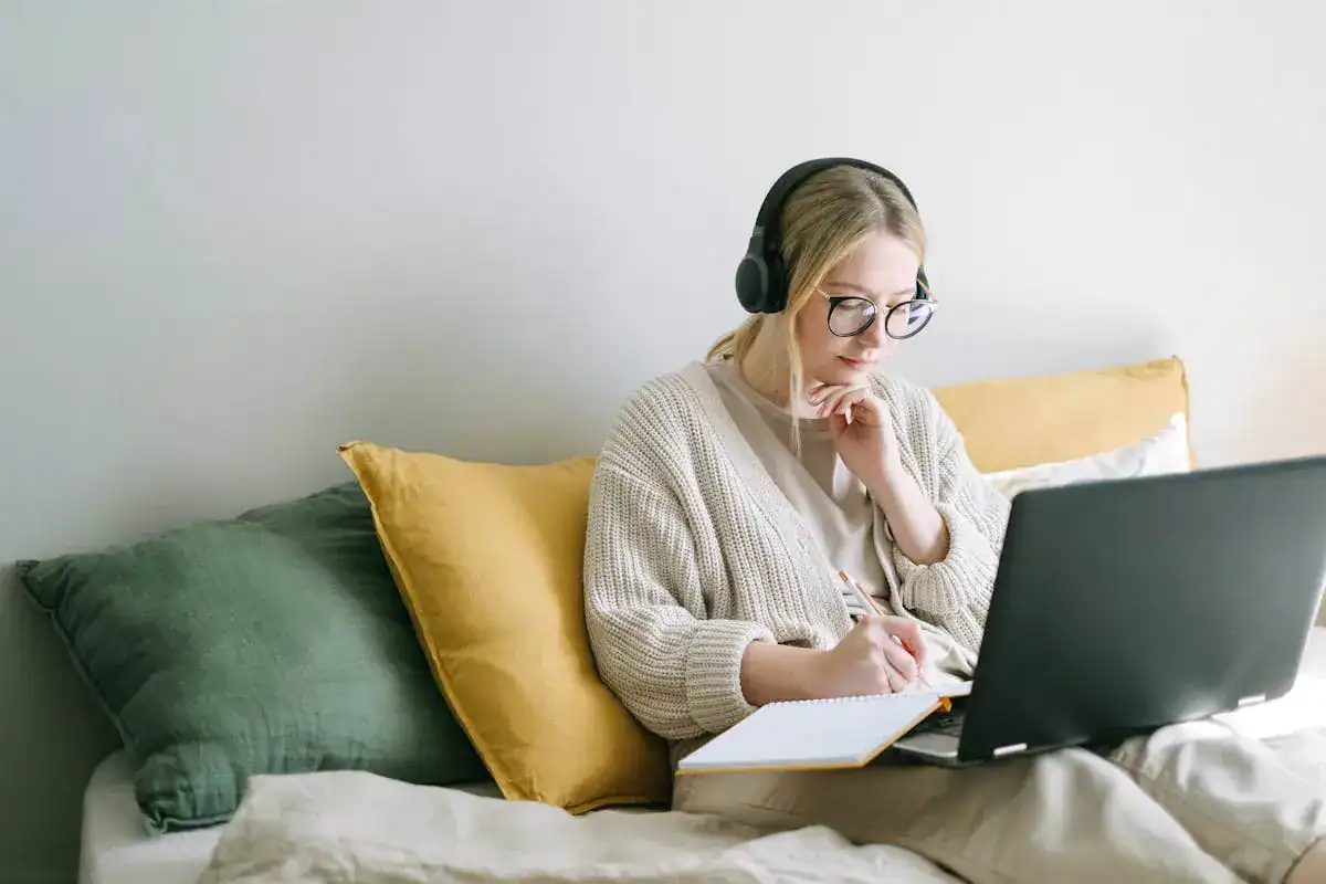 Woman working from her bed, computer and notebook with headphones on