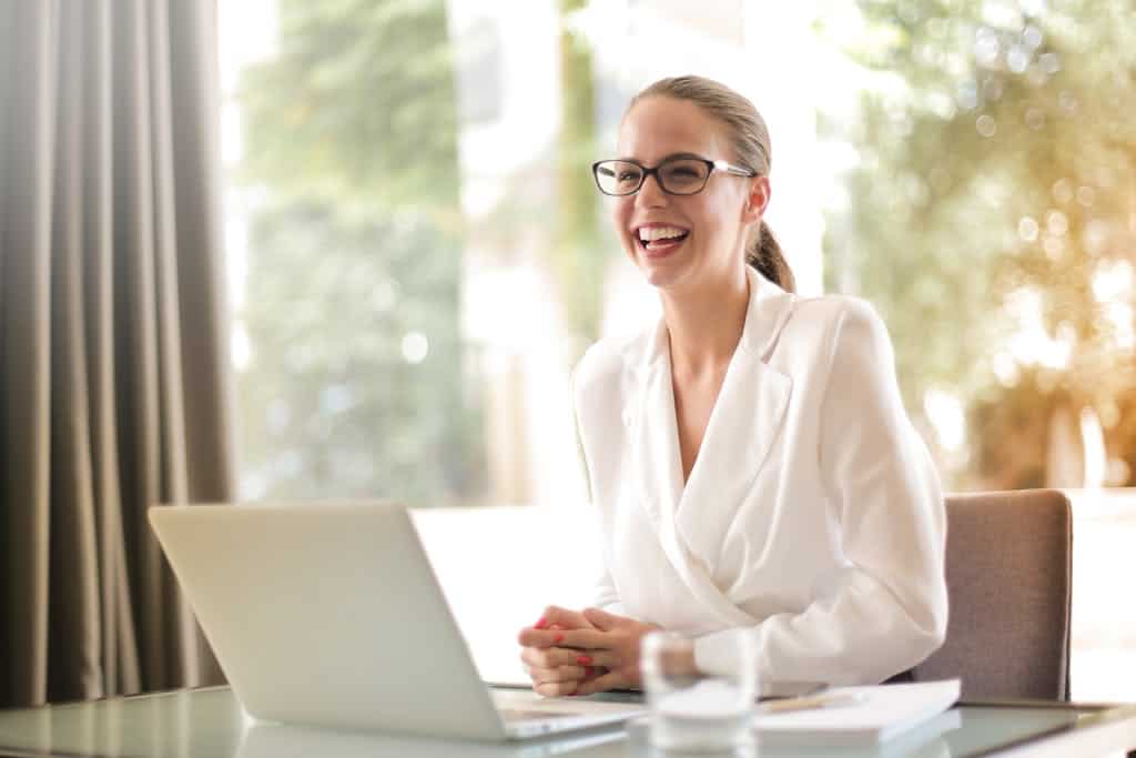Business Coaching Online. Cheerful businesswoman in glasses working on a laptop, in a bright and modern office setting.