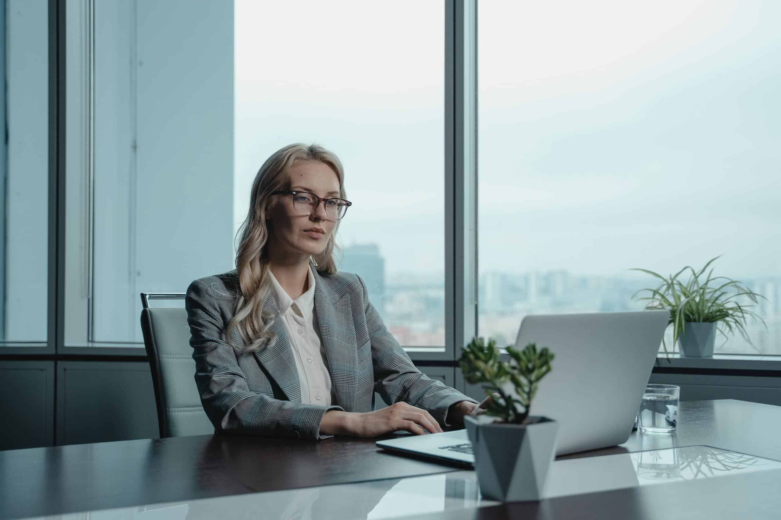 A confident businesswoman in a grey suit working on a laptop in a modern office setting. Fraactional CMO