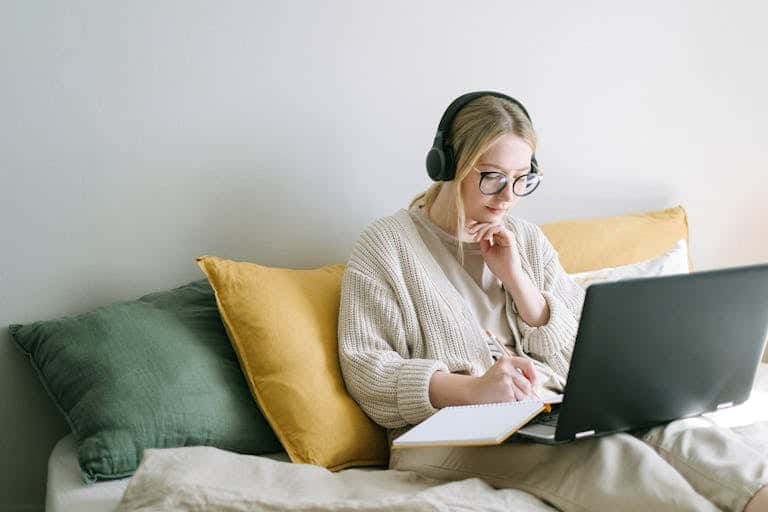 A focused woman in glasses and headphones works on a laptop from a cozy bed. Fractional CMO