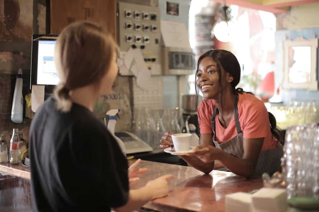 Smiling African American waitress wearing casual t shirt and apron standing at counter in cozy coffee shop with female customer while serving hot drink and looking at each other Client Journey Branding