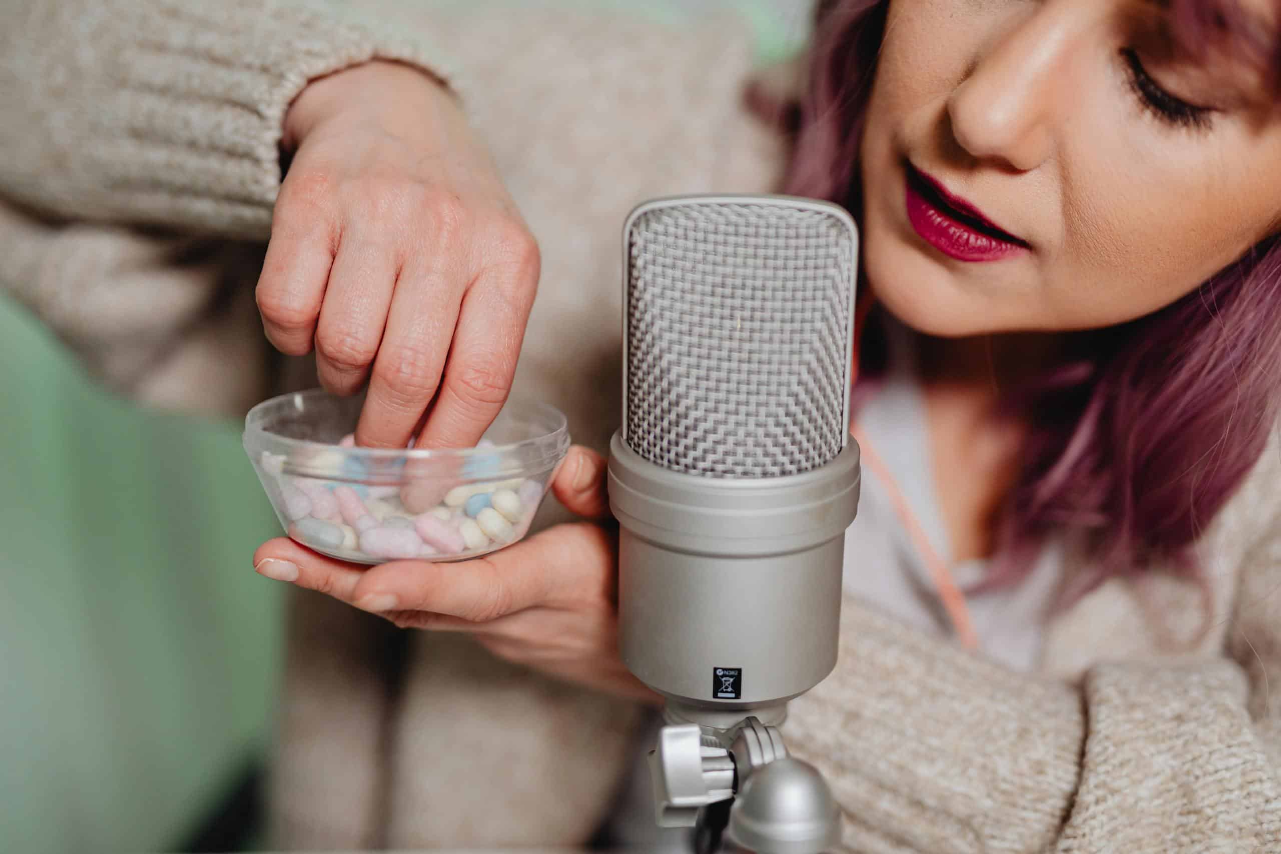 Woman in studio setting recording vlog, holding pills near microphone for ASMR content.