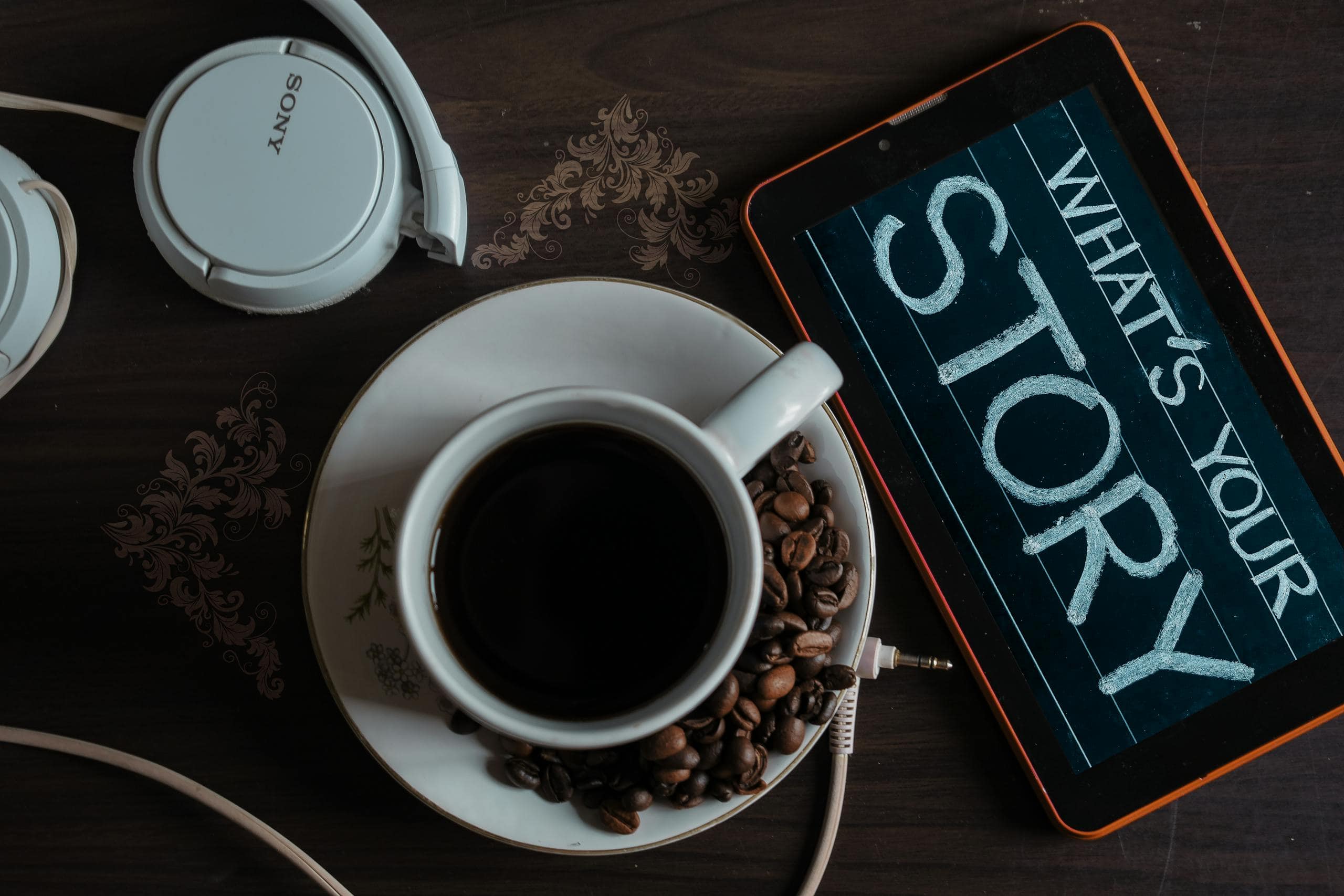 Flat lay of coffee cup, headphones, and tablet with 'What's Your Story' on screen. Digital Brand Storytelling