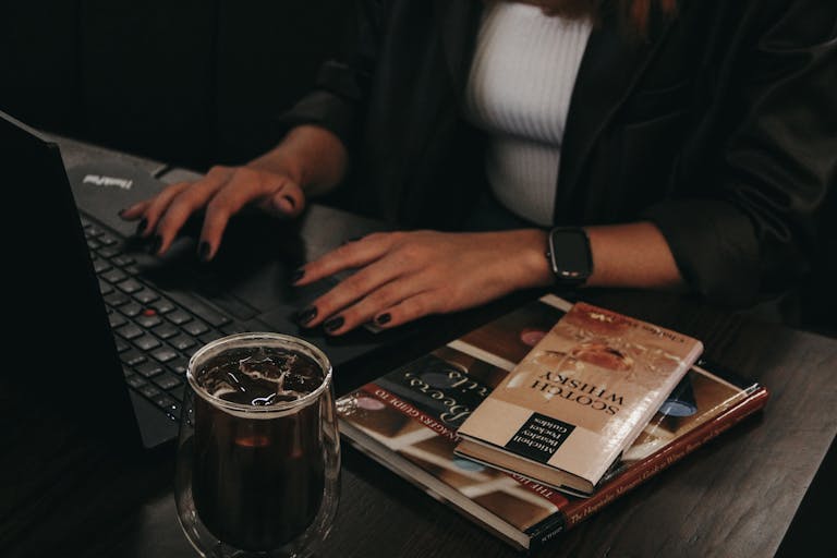 A woman typing on a laptop with coffee and books on a wooden table.