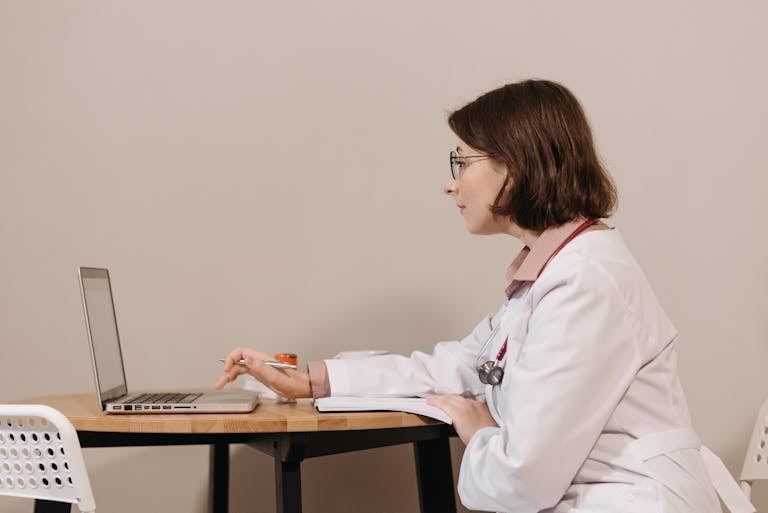 Side profile of a female doctor in a white coat working on a laptop at a desk.