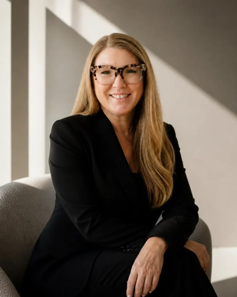 Smiling woman with long blond hair and tortoiseshell glasses sits on a grey sofa in a sunlit room, wearing a black blazer.